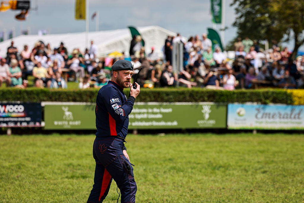 Having made safe the landing zone, Flight Lieutenant Adam Foster, 2IC RAF Falcons, talks to the team in the air via a handheld radio in front of a seated crowd.